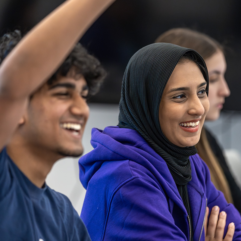 Person seated at a conference holding a microphone and a smartphone, with other attendees sitting nearby.